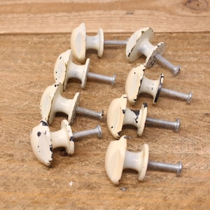 May include: Eight cream-coloured, oval cabinet knobs with distressed black detailing. Each knob is attached to a silver screw. The knobs are arranged on a wooden surface, suggesting a vintage or antique aesthetic.