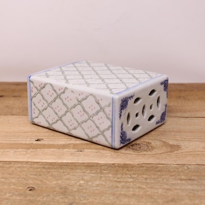 May include: A rectangular ceramic incense burner with a white base, adorned with a repeating pattern of green and pink floral motifs. The edges are trimmed with blue floral designs and the sides feature decorative cut-out holes.