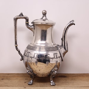 May include: A silver teapot with a geometric design and three lion paw feet. The teapot has a lid with a knob on top.