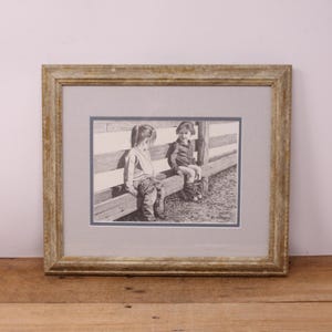 May include: A framed black and white pencil drawing of two children seated on a wooden fence. The artwork is matted and framed in a distressed gold-toned wooden frame. The drawing depicts the children in casual clothing.
