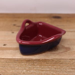 May include: A small, triangular ceramic dish with a handle. The interior and handle are a deep burgundy colour, while the exterior base is a contrasting navy blue. The dish sits on a wooden surface.