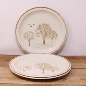 May include: Two cream-coloured ceramic plates with a brown rim and a tree design. The plates feature a landscape with bare trees and birds. The plates are stacked on a wooden surface.