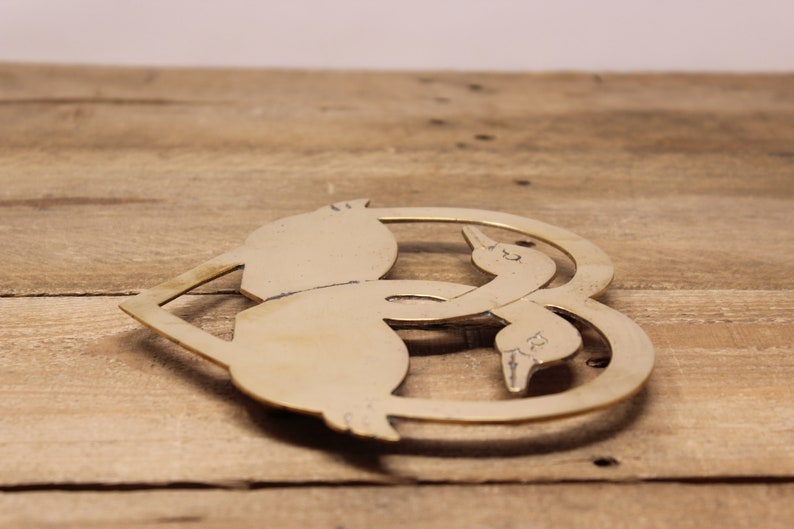 May include: A brass heart-shaped hot pad featuring two stylised geese intertwined. The geese are facing each other and their necks are curved to form the heart shape.