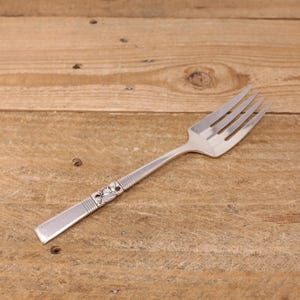 May include: A silver fork with four prongs, set on a wooden surface. The handle has a decorative floral pattern. The fork is angled, highlighting its polished finish and elegant design.
