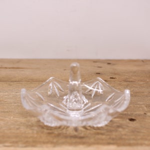 May include: A clear glass ring holder with a decorative design. The holder has a small handle on top.