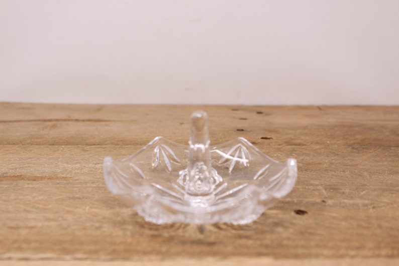 May include: A clear glass ring holder with a decorative design. The holder has a small handle on top.