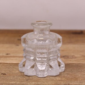 May include: A clear, vintage glass perfume bottle with a geometric design. The bottle has a round base and a short, cylindrical neck. The body of the bottle is faceted, creating a diamond-like pattern. The bottle is empty and sits on a wooden surface.