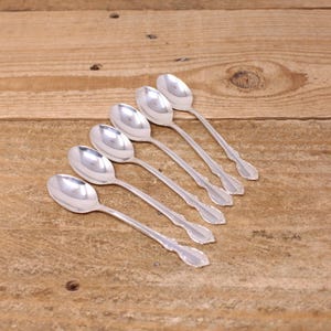 May include: Six silver spoons with ornate handle detailing are arranged diagonally on a wooden surface. The spoons have a reflective, polished finish, suggesting they are made of stainless steel or a similar metal. The spoons are likely for serving or eating.