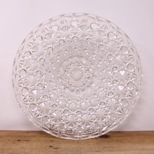 May include: A clear, circular glass serving plate featuring an intricate starburst design. The plate has a raised edge and a central pattern. The detailed design creates a sparkling effect, suitable for serving food or as a decorative item. The diameter is approximately 30 cm.