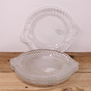 May include: Clear glass plates with a fish design. The plates have a textured surface and a fish-shaped outline, including fins and a tail. The plates are stacked on a wooden surface.