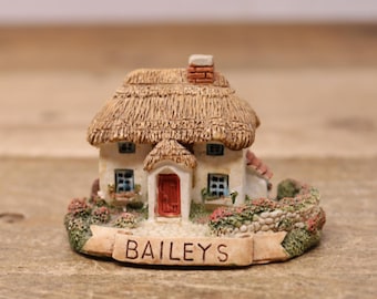 May include: A miniature thatched-roof cottage with a red door and white walls. The cottage is surrounded by green foliage and a stone wall. A banner with the text "BAILEYS" is displayed in front of the cottage.