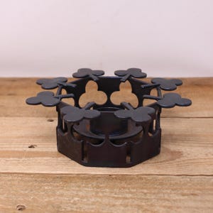 May include: A dark metal candle holder with a clover-shaped design. The candle holder has a circular base with a series of cut-out shapes and clover-shaped accents extending outwards. The holder is sitting on a wooden surface.