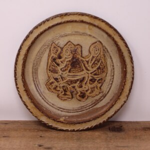 May include: A round, beige ceramic plate with a raised, textured rim. The plate features a central embossed design of three figures in a circle. The figures are in a dark brown color, and the plate has a rustic, handmade appearance.