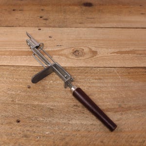 May include: A vintage metal and wood can opener with a brown handle. The opener has a lever and a blade for opening cans.