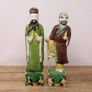 May include: Two ceramic figurines of Asian figures. One figure wears a green robe and holds two yellow objects. The other figure wears a brown robe and leans on a yellow chair. Both figures stand on decorative pedestals with green and yellow accents.