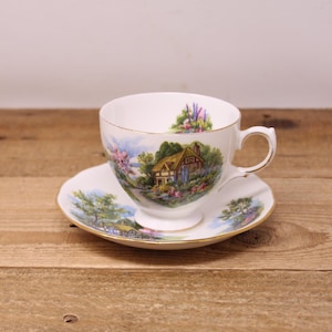 May include: White teacup and saucer set with a gold rim. The teacup and saucer feature a scenic landscape design with a cottage, trees, and flowers. The teacup is resting on the saucer on a wooden surface.