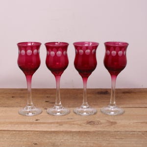 May include: Four vintage ruby red cordial glasses with white polka dot detailing around the rim. The glasses have a clear stem and base, and are arranged on a wooden surface.