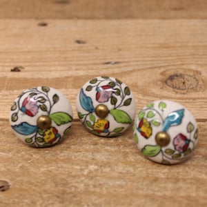 Peut inclure: Trois boutons de meuble en céramique blanche avec des motifs floraux. Chaque bouton a une base dorée et est orné de fleurs colorées, de feuilles vertes et d'accents bleus. Les boutons sont posés sur un fond en bois.
