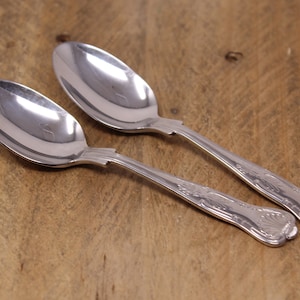 May include: Two polished silver spoons with decorative handles rest on a wooden surface. The spoons have a traditional design, featuring a rounded bowl and an ornate pattern on the handle.