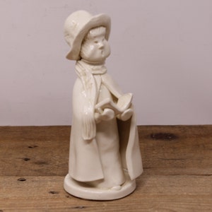 May include: White ceramic figurine of a person wearing a hat and a long coat, holding a book. The figurine is standing on a round base.
