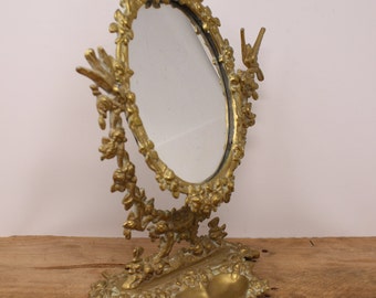 Può includere: Uno specchio da toeletta ovale ornato, color oro, con una base decorativa. Lo specchio è circondato da un motivo floreale e ha un piccolo piatto sulla base.