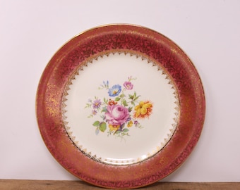 May include: A vintage floral dinner plate with a white center and a pink rim with gold accents. The center features a bouquet of pink, yellow, and blue flowers.