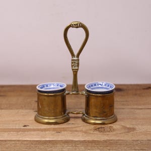 May include: A vintage brass condiment holder with two cylindrical cups. Each cup has a small, white and blue patterned dish inside. The holder has a handle at the top. The item is on a wooden surface.