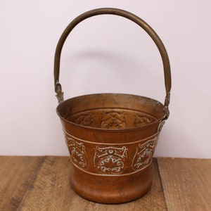 May include: A copper bucket with a brass handle. The bucket has a white, decorative design around the top and bottom.