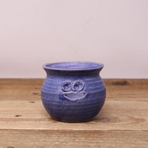 May include: A small, round, blue ceramic pot with a smiling face carved into the front. The pot has a textured surface and sits on a wooden surface. The pot is likely used for holding small plants or other decorative items.