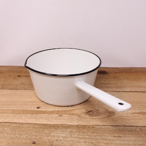 Peut inclure: Une casserole en émail blanc avec un bord noir et une poignée unique. Le récipient présente un petit bec verseur sur le côté gauche et un trou circulaire à l'extrémité de la poignée pour le suspendre. La finition est lisse et brillante.