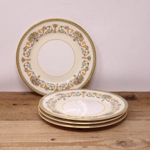 May include: A set of fine china plates with a cream-colored background and a decorative floral border in shades of blue, yellow, and red. The plates have a gold rim and are stacked on a wooden surface.