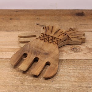 May include: Two wooden salad servers with a hand-like design. The servers are made of light brown wood and feature a decorative carved pattern on the handle. The tines are curved and spaced apart. The servers are resting on a wooden surface.