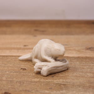 May include: An off-white beaver figurine, resting on a log. The sculpture has detailed fur and is in a relaxed pose. The background is a wooden surface, providing a natural setting for the animal.