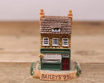 May include: A miniature model of a two-story building with a brown roof, yellow brick facade, and a green storefront. The storefront has a sign that reads "The Corner Shop" and a sign that says "Lounge". The base of the model is decorated with green foliage and reads "Bailey's '95".