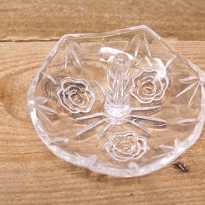 May include: A clear glass ring holder with a raised center post and a decorative etched design of roses.