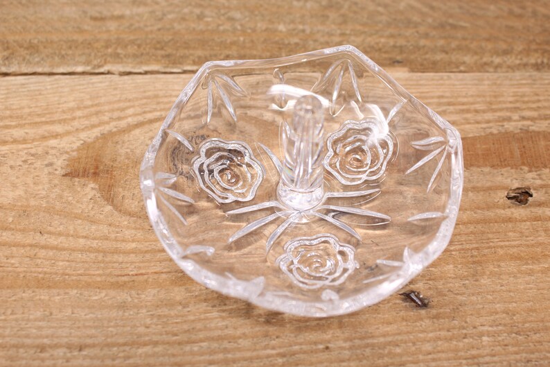 May include: A clear glass ring holder with a raised center post and a decorative etched design of roses.