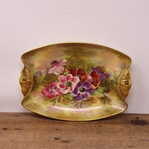May include: An antique, oval decorative dish with a gold rim and handles. The dish features a hand-painted floral design with pink, purple, and red flowers. The background has a soft, green and gold colour palette.