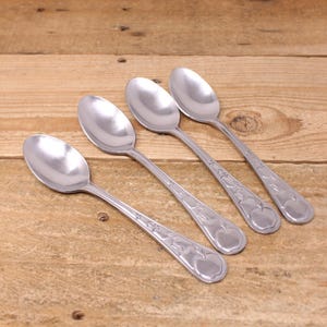May include: Four stainless steel spoons with an apple design on the handles, arranged diagonally on a wooden surface. The spoons have a shiny, reflective surface, and the apple design is etched into the metal.