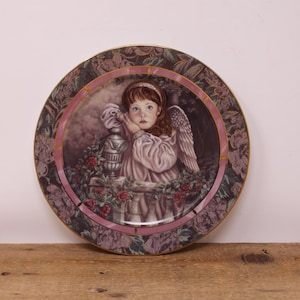 May include: Decorative plate featuring a young girl with angel wings, resting her chin on a decorative urn. The plate has a floral border with gold trim and a pink inner ring. The girl is wearing a pink dress and a headband.