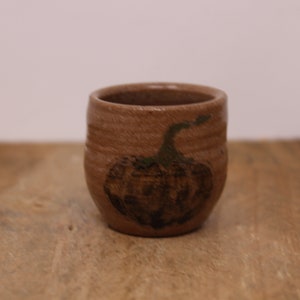 May include: A small, handmade brown ceramic cup with a pumpkin design. The pumpkin is dark brown with green accents. The cup is cylindrical with a slightly flared rim and sits on a wooden surface.