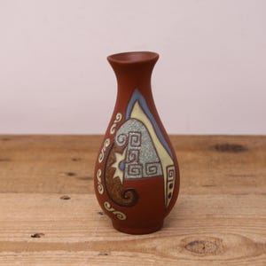 May include: A small, brown ceramic vase with a geometric design in shades of blue, green, and yellow. The vase has a rounded body and a narrow neck.
