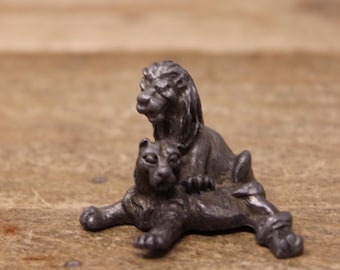 May include: A small pewter figurine of two lions, one sitting on top of the other. The lion on top is looking to the left, while the lion on the bottom is looking to the right.
