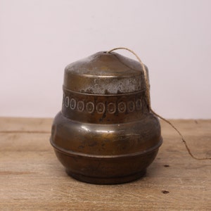 May include: A vintage brass container with a lid and a decorative band of circles. A string is attached to the lid. The container is sitting on a wooden surface. The container has a tarnished appearance.