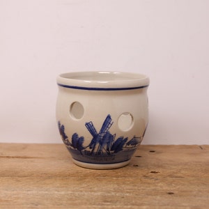 May include: A white ceramic pot with blue and white Delftware designs of a windmill and houses. The pot has several holes around the sides.