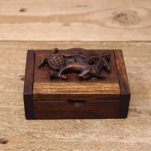 May include: A small, square wooden box with a hinged lid. The box is stained a dark brown color. The top of the box features carved relief images of a dolphin, starfish, turtle, and sand dollar. The box is sitting on a wooden surface.