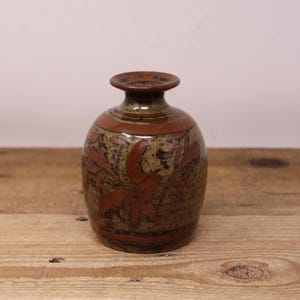 May include: A small, handcrafted ceramic vase with a rounded body and a narrow neck. The vase features a brown and olive green colour scheme with reddish-brown abstract designs. The vase sits on a wooden surface.