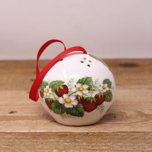 May include: A white ceramic ornament with a red ribbon. The ornament is decorated with a pattern of strawberries, white flowers, and green leaves. It has several small holes at the top.