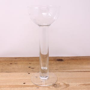 May include: A clear glass vase with a round base and a long, slender stem. The vase has a wide, rounded top. The glass is transparent, allowing light to pass through. The vase is displayed on a wooden surface.