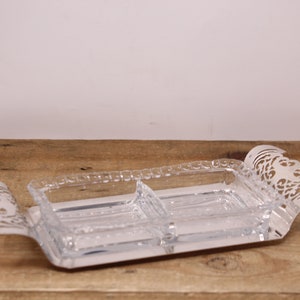 May include: A clear glass serving dish with two compartments, set on a silver tray with an ornate design. The tray has a decorative, cut-out pattern around the edges.