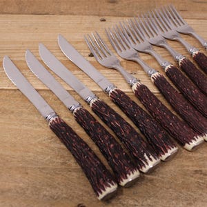 May include: A collection of stainless steel knives and forks. The cutlery features brown and cream-coloured antler handles. The knives have serrated blades. The silverware is displayed on a wooden surface.